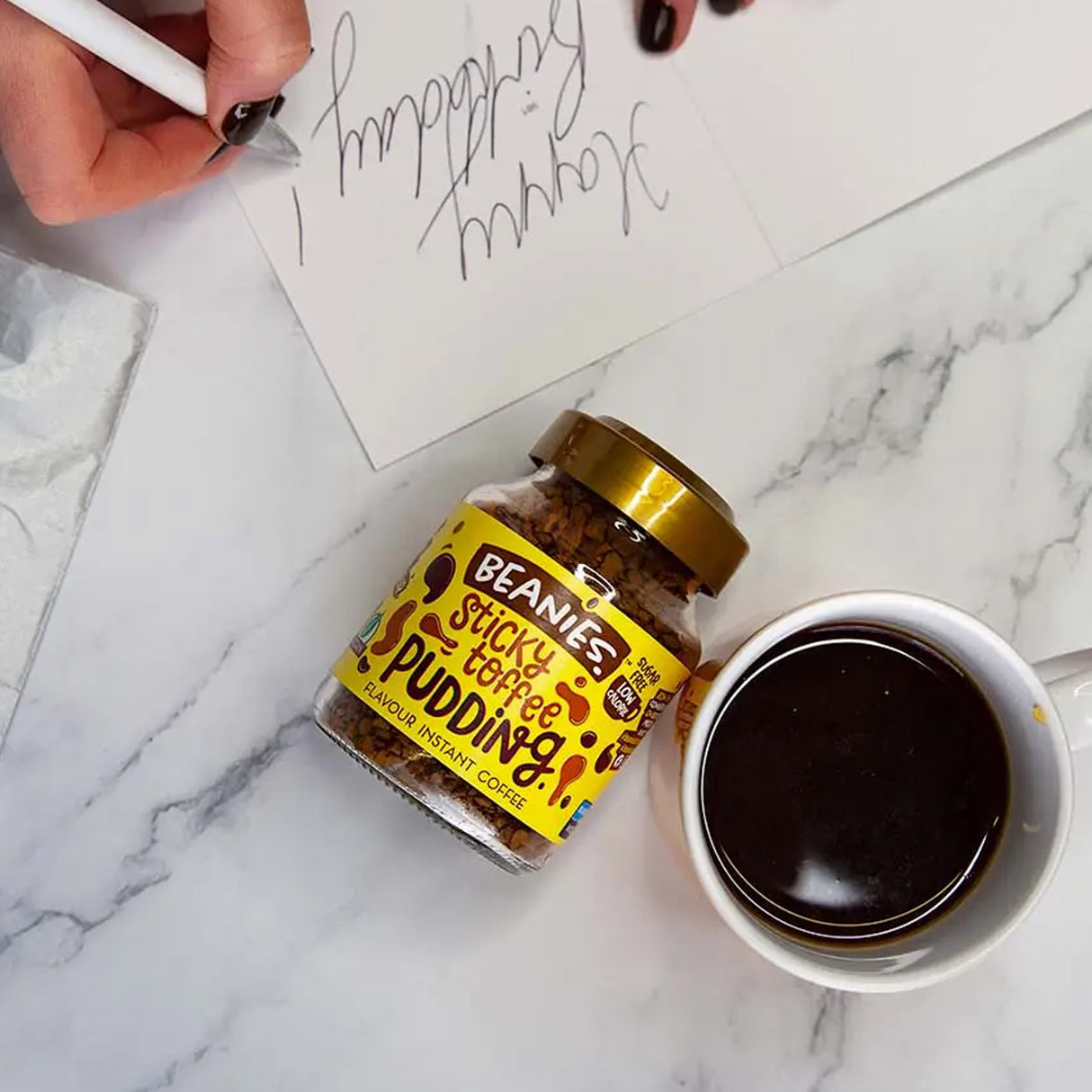 Jar of Beanies Sticky Coffee Pudding on a marble surface with a cup of coffee and a hand writing.