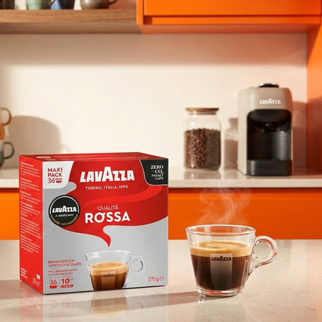 A box of Lavazza Rossa coffee capsules sits on a kitchen counter next to a glass mug filled with espresso, with a coffee machine and jars in the background.