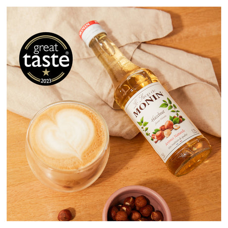 Monin bottle of hazelnut syrup next to a cup of coffee on a wooden surface with 'Great Taste' award logo.