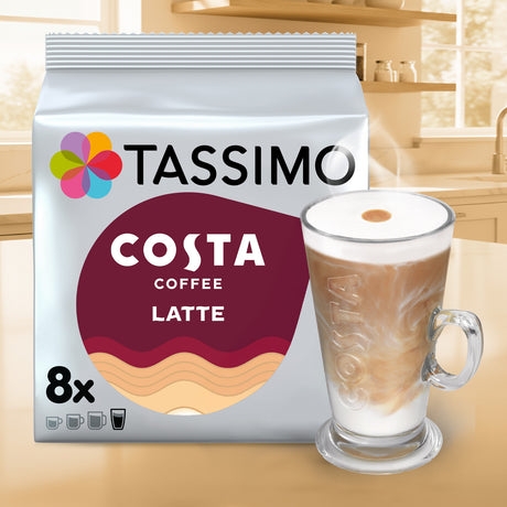 A package of Tassimo Costa Coffee Latte stands on a countertop next to a glass mug filled with a layered coffee and milk latte. The bag is white with a large burgundy and beige oval label and the Tassimo logo.