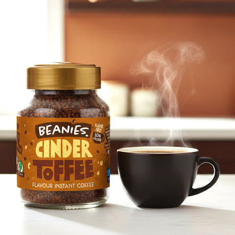 Jar of Beanies Cinder Toffee instant coffee with a steaming cup on a kitchen counter.