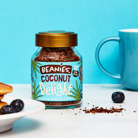 Jar of Beanies Coconut Delight instant coffee with a blue mug and blueberries on a white surface.
