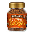 Jar of Beanies Creamy Caramel instant coffee on a wooden table with a steaming cup of coffee.