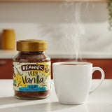 Jar of Beanies Very Vanilla Decaf instant coffee with a steaming white mug on a kitchen counter.
