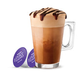 A tall clear glass mug filled with a mocha latte topped with whipped cream and chocolate drizzle, shown on a white background with two purple Nespresso Dolce Gusto coffee capsule packs beside it.