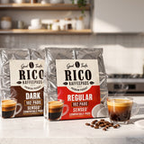 Two bags of RICO Kaffepads coffee filter pods, one Dark and one Regular, displayed on a kitchen counter with espresso cups and coffee beans.