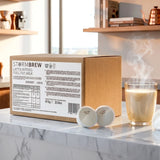 Box of Storm Brew milk with two milk pods and a glass of steaming milk on a marble surface.