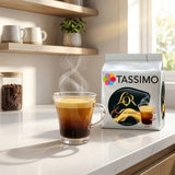 Tassimo coffee pod packaging with a steaming glass of coffee on a kitchen counter.