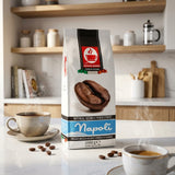 A bag of Napoli espresso coffee stands on a kitchen counter with two cups of coffee and scattered coffee beans in a warm, blurred kitchen setting.